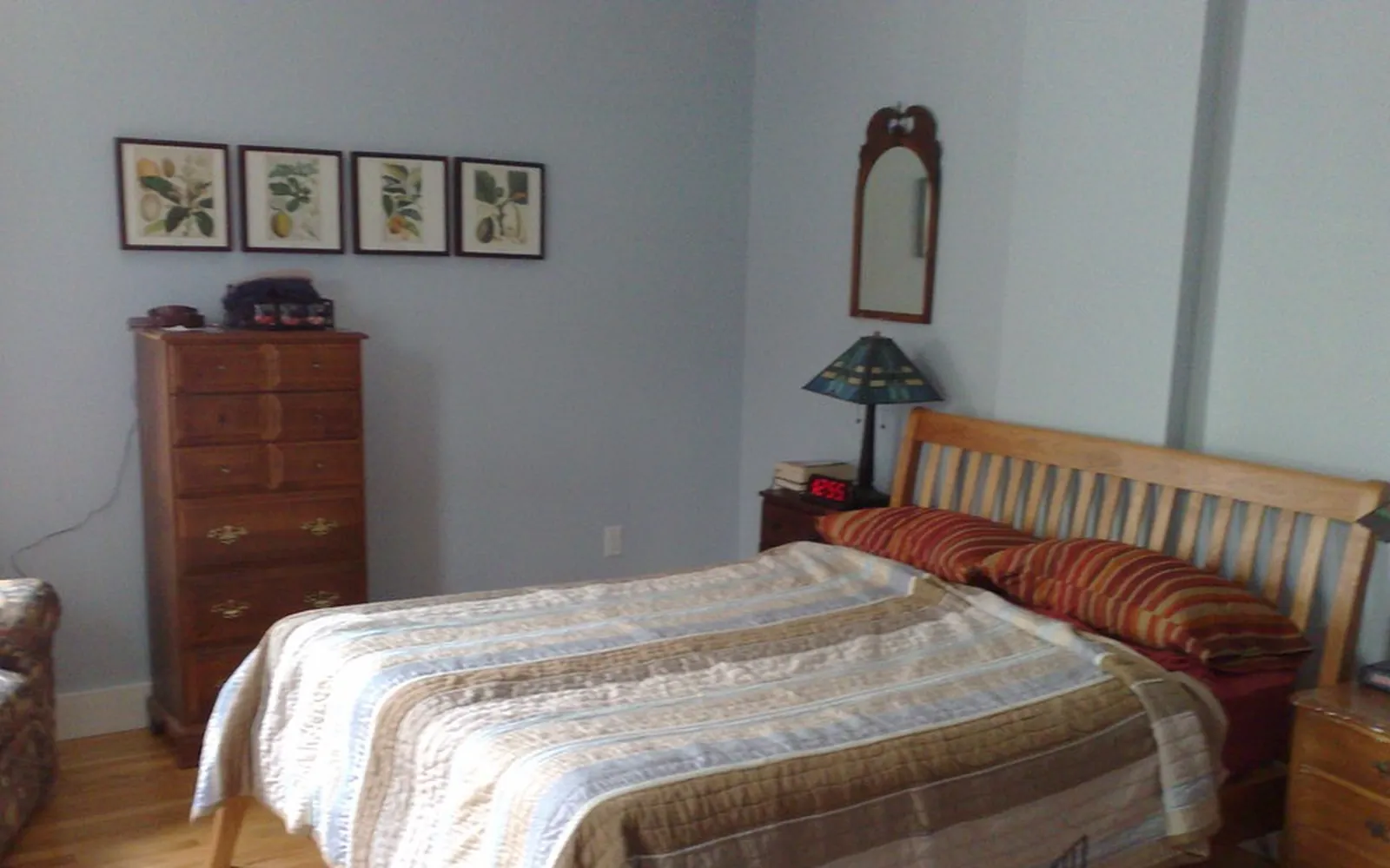 A wooden double bed with a striped quilt and red pillows in a plain bedroom with a chest of drawers